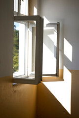 Open white-framed window in a yellow and white painted room with sunlight casting geometric shadows on the wall. Green foliage is visible through the glass, creating a peaceful and airy indoor atmosph