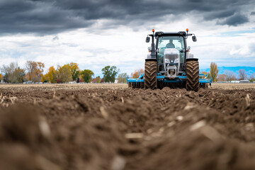 Fototapeta premium Tractor with plow turning dark soil under cloudy autumn sky, 