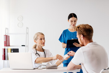 Obraz premium Doctor in white coat consulting with male patient, while nurse assists with medical supplies in modern clinic, showcasing healthcare teamwork and patient care environment