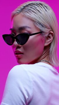 Fashion portrait of a confident young Asian woman with trendy blonde hair and sunglasses looking over her shoulder on a vibrant pink background with neon lighting