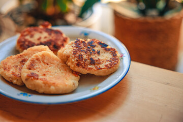 A cozy homemade breakfast featuring traditional syrniki (cottage cheese pancakes) served with butter and sausage. Set in a warm, rustic home environment with soft morning light, this comforting meal e