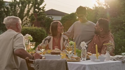 Medium shot of young boy running up to dinner table outdoors and embracing mom and dad enjoying cozy family gathering in hazy Summer glow with lens flare effect - Powered by Adobe