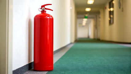 Bright red fire extinguisher stands ready for use in a long dimly lit hotel hallway with green carpeting and white walls