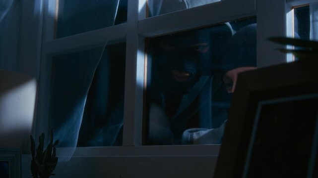 Close up of two criminal partners in black balaclavas looking through window while discussing plan of break in