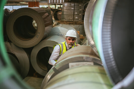 Senior factory manager in safety helmet walking to inspect large metal sheet rolls at production facility, demonstrating leadership, quality control, and high industry professionalism.