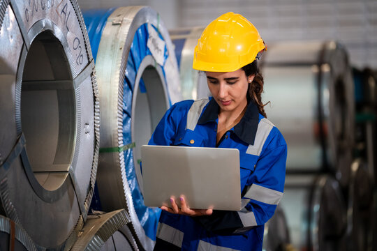 Female industrial engineer in safety helmet using laptop to inspect large metal coil rolls in manufacturing factory, ensuring quality control and efficient production process.