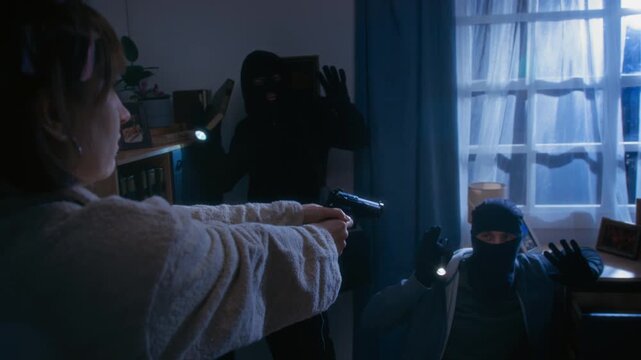 Over shoulder view of woman in bathrobe pointing at burglars with gun while threatening during unlawful invasion