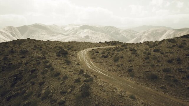 A winding dirt road cuts through the rugged terrain of the Pamir Mountains in Tajikistan, surrounded by rocky hills and dramatic mountain landscapes under a cloudy sky.