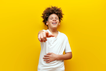 cheerful curly african american boy making fun and joking and pointing at camera on yellow isolated background, teenager in white t-shirt mocking and showing funny © Богдан Маліцький