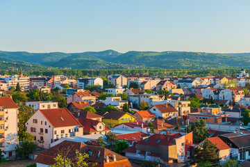 Panoramic view of a sunlit Novi Sad in Serbia town with a mix of modern and traditional houses. Rooftops stretch toward the horizon with green hills in the background.