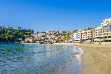  Mali Beach in Ulcinj, Montenegro, with golden sand and calm Adriatic waters. Colorful hillside buildings and the historic fortress overlook the vibrant waterfront under a clear blue sky.