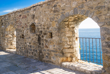  Stone wall with arched openings in Ulcinj’s Old Town, Montenegro, overlooking the Adriatic Sea. Sunlight illuminates the historic masonry, revealing a scenic coastal view through the arches.