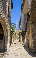 Narrow stone alley with steep steps in Ulcinj’s Old Town, Montenegro, flanked by historic stone houses. Sunlight brightens the charming medieval passage under a vivid blue sky.