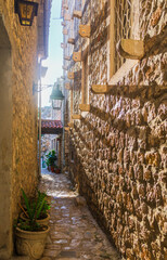 Narrow stone alley in Ulcinj’s Old Town, Montenegro, adorned with potted plants and wrought-iron lanterns. Warm sunlight casts shadows along the textured medieval walls.