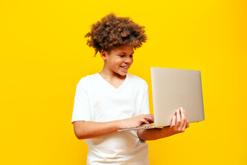 cheerful african american boy in white t-shirt using laptop on yellow isolated background and smiling, curly schoolboy typing on computer online and looking at camera