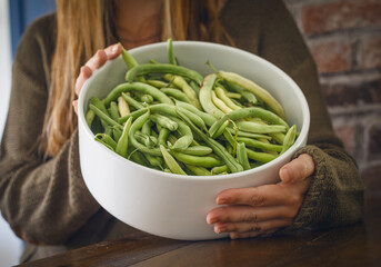 The woman holds a large white bowl filled with fresh green and yellow beans. He is wearing a cozy swamp sweater, creating a warm and homey atmosphere.
