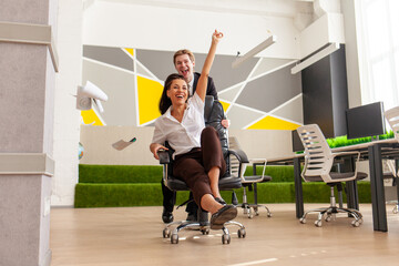 Fototapeta premium office workers in business suits having fun and riding on chairs with raised hands, african american woman with european young man celebrating success and rejoicing in victory at work