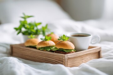 A wooden tray with mini burgers and coffee on a white bed sheet with a plant in the background