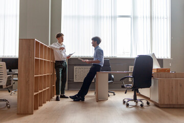 young male boss in business clothes sitting in office at table and talking with colleague at work, entrepreneur and his partner smiling and discussing report and plan