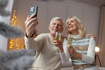 elderly couple of seniors in sweaters celebrate the New Year at home drinking champagne and taking selfies on smartphone, grandfather and grandmother communicate via video call on Christmas