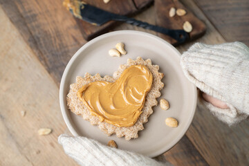 Hands covered with a cozy wool sweater holding a plate with  heart shaped whole wheat bread smothered with creamy peanut butter.  Comfort food.