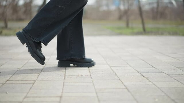 Side view close-up of person in black boots and dark jeans walking on paved surface in cold outdoor park, one foot slightly raised in mid-step, showcasing motion, balance, and calm winter atmosphere