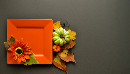 Autumn table setting with orange square plate decorated with artificial sunflower and green leaves. Nearby are decorative mini pumpkins, autumn leaves and plastic black spiders on a dark background. 