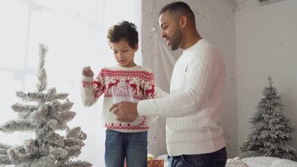 African American man with his son in New Year's clothes decorate the tree with toys and celebrate Christmas at home, father helps the boy prepare the New Year's atmosphere - Powered by Adobe
