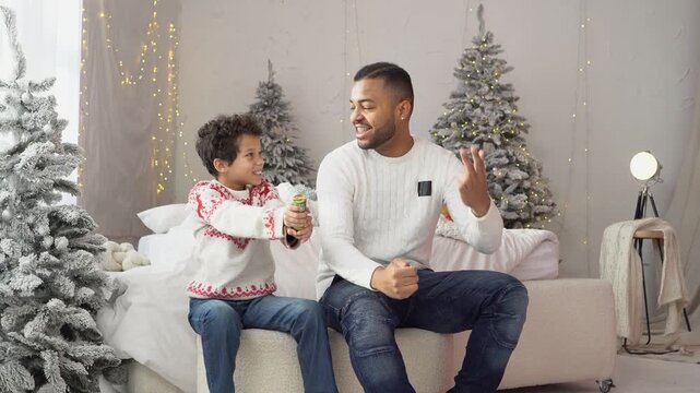 African American father and son blowing confetti on New Year's Eve as the clock counts down, boy holding cracker and rejoicing, man and teenager celebrating Christmas together at home