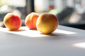 Three peaches are lined up on a white surface, illuminated by sunlight