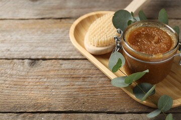 Natural body scrub in glass jar, brush and eucalyptus branch on wooden table, closeup. Space for text