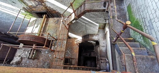 Derelict industrial rusted stairwell. Rusty corroded metal railings, factory cogs and machinery. Downward spiral steps. Abandoned decaying industry building staircase. Stairs atmosphere looking down.