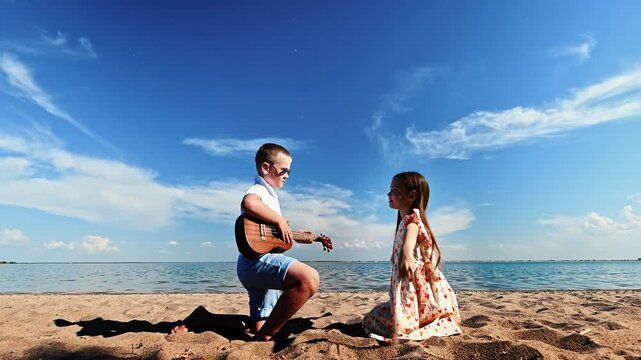 Two small children relax on sandy beach near lake on warm summer day: little boy plays guitar for beautiful girl against background of blue clear sky and water. Leisure time activity outdoors.