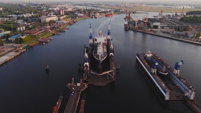 A shipyard in Riga, Latvia, features a large vessel docked for maintenance, cranes in motion, smaller boats, and industrial buildings by a river. - Powered by Adobe