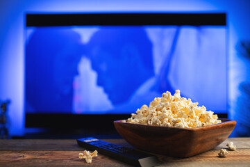 A wooden bowl of popcorn and remote control in the background the TV works. Evening cozy watching a movie or TV series at home