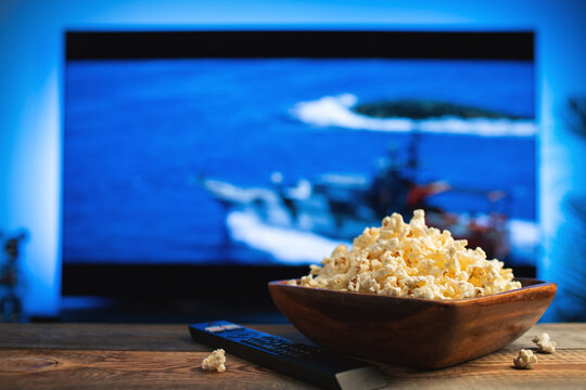 A wooden bowl of popcorn and remote control in the background the TV works. Evening cozy watching a movie or TV series at home - Powered by Adobe