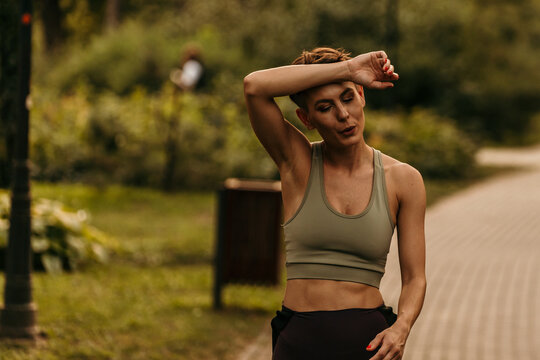 Tired sportswoman wiping sweat from forehead after training in park - Powered by Adobe