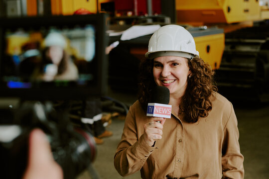 Caucasian young adult woman wearing hard hat smiling and holding microphone with TV news logo while being filmed in industrial setting, construction equipment visible in background