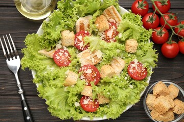 Tasty salad with croutons, tomatoes, cheese and chicken on black wooden table, flat lay