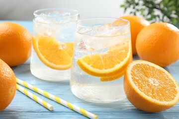 Sparkling water with orange slices in glasses, fresh fruits and straws on light blue wooden table, closeup