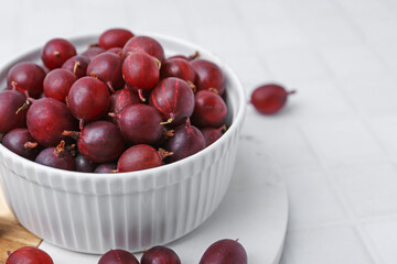 Ripe red gooseberries in bowl on white tiled table, closeup