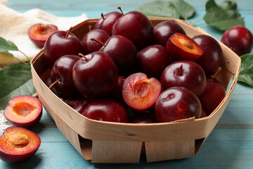 Whole and cut cherry plums in basket on light blue wooden table, closeup
