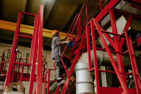 Unrecognizable woman wearing safety helmet climbing industrial metal stairs in factory setting, holding railing with gloved hands, appearing focused on maintenance or inspection task