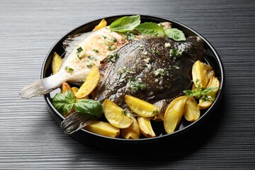 Tasty roasted flounder fish with herbs and potato on black table, closeup. Homemade seafood dish
