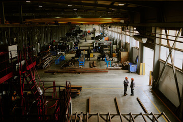 Obraz premium Two middle aged Caucasian women standing and talking in large industrial factory workshop, surrounded by heavy machinery and metal materials, wearing protective helmets and workwear