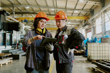 Caucasian young adult woman and Caucasian middle aged woman wearing safety helmets standing in factory, discussing project while using digital tablet, industrial equipment in background