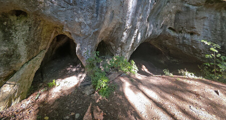 Wide view of a limestone cliff with three distinct cave entrances in the Bükk Mountains, Hungary, surrounded by forest vegetation.