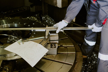 Unrecognizable woman measuring large metal component with caliper in industrial workshop, holding technical drawing on machinery, wearing protective gloves and work uniform