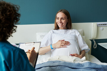 Caucasian young adult woman sitting in hospital bed smiling and holding pregnant belly while female...