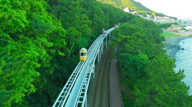 Aerial view of Sky Capsules traveling along an elevated railway track through a green forest, with a coastal town and ocean, in Busan, South Korea.